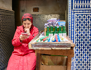 Woman using smartphone in Fez Medina while sitting by a stall by Marco Brivio