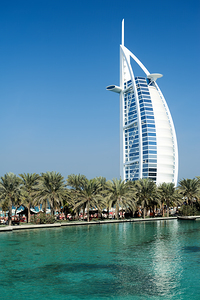 Burj Al Arab hotel stands tall by the water with palm trees by Marco Brivio