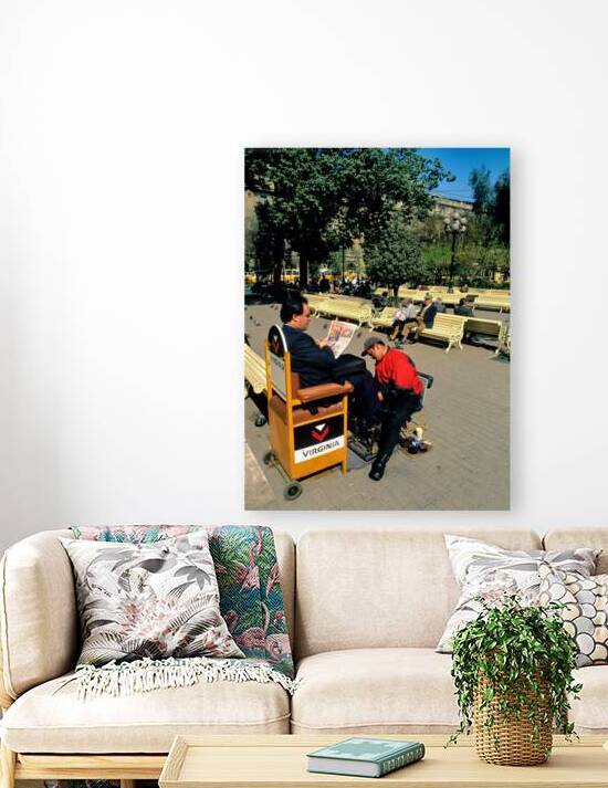 Man gets shoeshine in park while reading newspaper. by Marco Brivio
